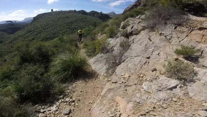 MTB Bike Station - Downhill Enduro - Santa Ponca Wanderer auf einem schmalen Weg durch grüne Hügel und felsige Landschaft.